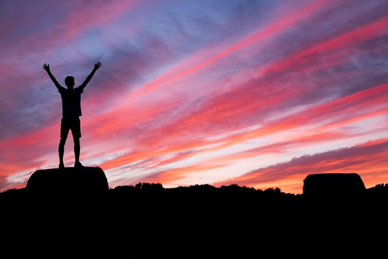 Bilde av en mørklagt person med hendene oppe i horisonten. Kraftig rød og blå himmel i bakgrunn. Kilde: unsplash.com