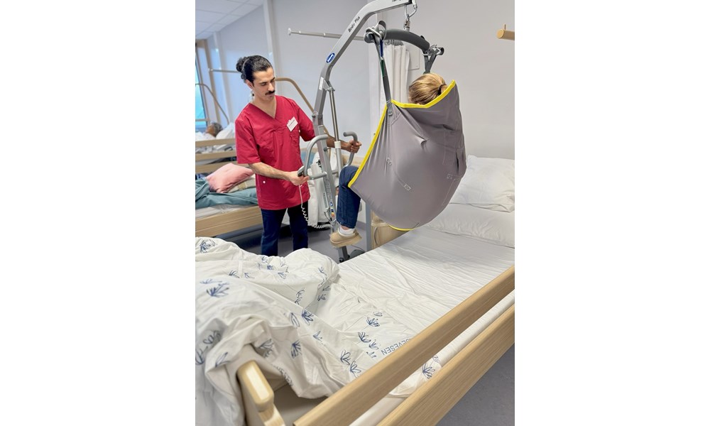 Personheis-bildet Bilde av en uniformert helsefagarbeider som heiser en medelev opp fra en sykehusseng med en mobil pasientheis