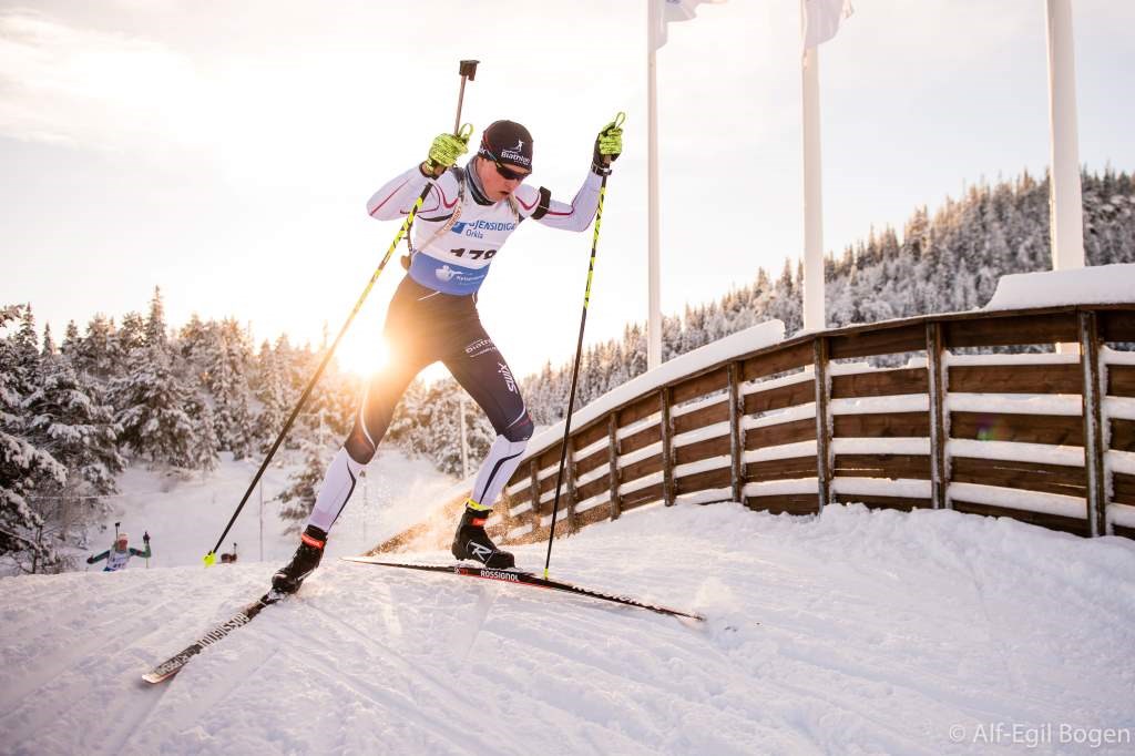 En skiskytter med gevær på ryggen som går på ski over en snødekt ski-bro med sola i ryggen.