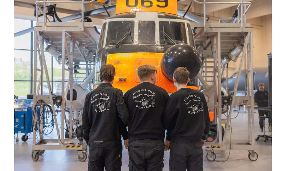 Bildet viser tre personer med ryggen til, som står i en flyhangar og ser på et stort helikopter. De er elever ved flyfag-linjen på Fosen videregående skol