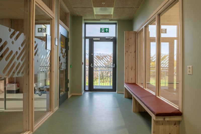 Bildet viser en korridor eller et venteareal preget av lyse trematerialer og store glassflater.