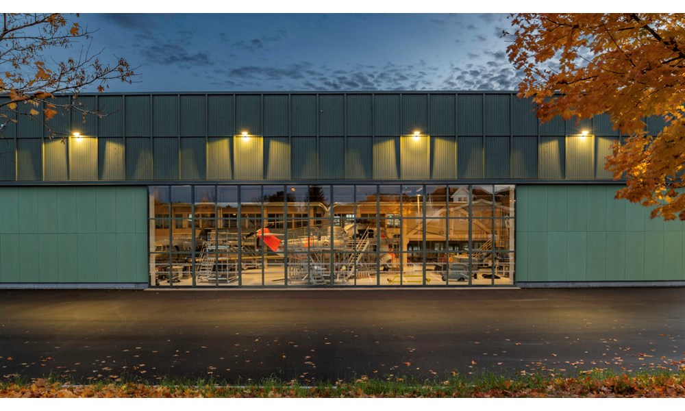 Dette bildet viser hangarbygget fra utsiden i skumringen, hvor de store glassflatene gir fullt innsyn til helikopteret og verkstedområdet på innsiden.