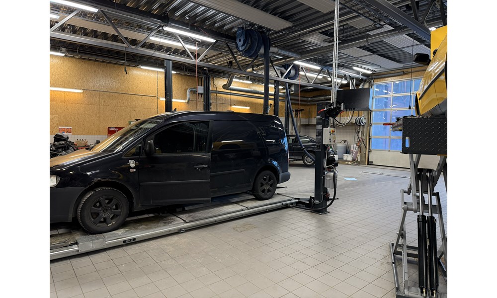 Midt i bildet står en svart varebil (ligner en Volkswagen Caddy) plassert på en kjørebaneløfter – en type heis som løfter bilen etter hjulene.