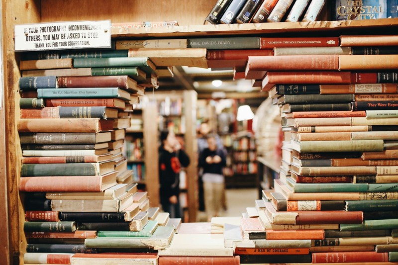 Bildet viser bøker stablet i en bokhylle. Bøkene er stablet slik at det er en åpen sirkel i midten, hvor en kan se gjennom og se mennesker i et bibliotek i bakgrunnen.