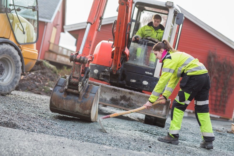 Ei jente graver i bakken med ei hakke, mens en gutt sitter i en gravemasking i bakgrunnen
